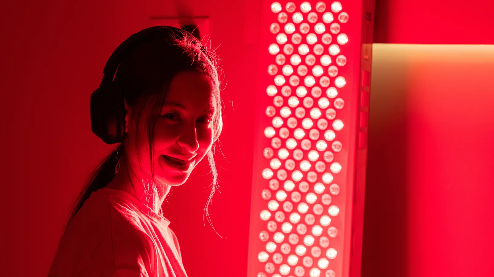 A woman standing beside a tall red light therapy panel in a home room, the panel throwing deep red light across the whole space.